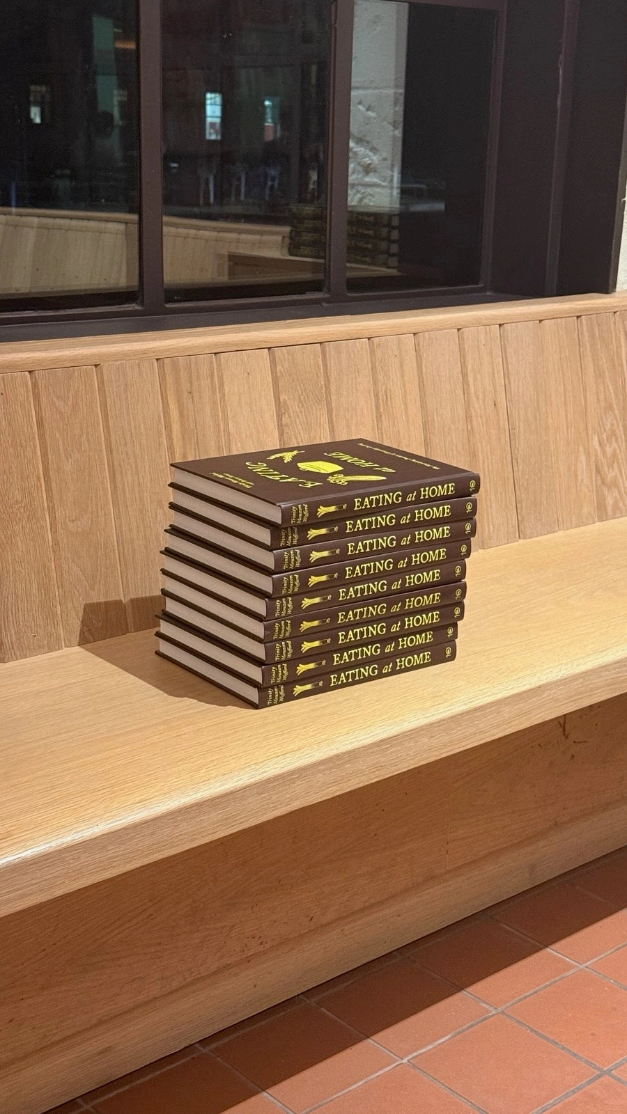 A stack of Eating at Home cookbooks on a wooden bench